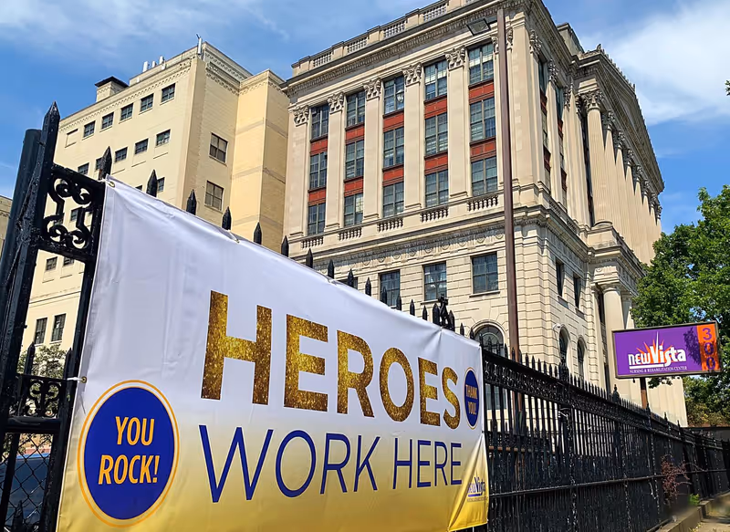 Exterior view of a large, multi-story classical building with ornate architectural details under a blue sky. In the foreground, a white banner with gold and blue text reads 'HEROES WORK HERE' and 'YOU ROCK!'. A purple sign on the fence reads 'New Vista Nursing & Rehabilitation Center'.
