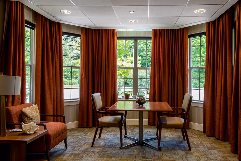 A cozy sitting area with a wooden table and two chairs positioned in front of large windows with brown curtains. There is an orange armchair with a patterned cushion and a side table with a lamp and decorative items. Outside the windows, green trees and bushes are visible.