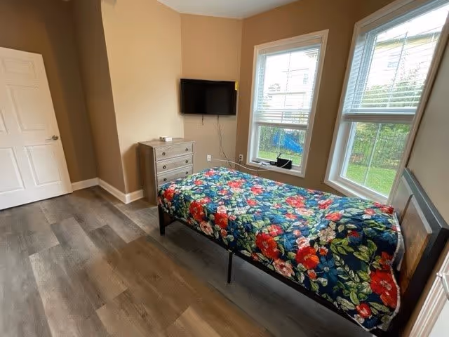 A small bedroom with a single bed covered in a colorful floral blanket. There is a chest of drawers against the wall with a flat-screen TV mounted above it. Two large windows let in natural light and show a view of a green outdoor area. The room has wood flooring and beige walls.