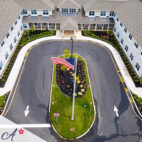Aerial view of the entrance driveway of a large assisted living facility building with a U-shaped driveway surrounding a landscaped island featuring an American flag on a flagpole. The building has multiple windows and a covered entrance with columns. The driveway has directional arrows and a stop sign.