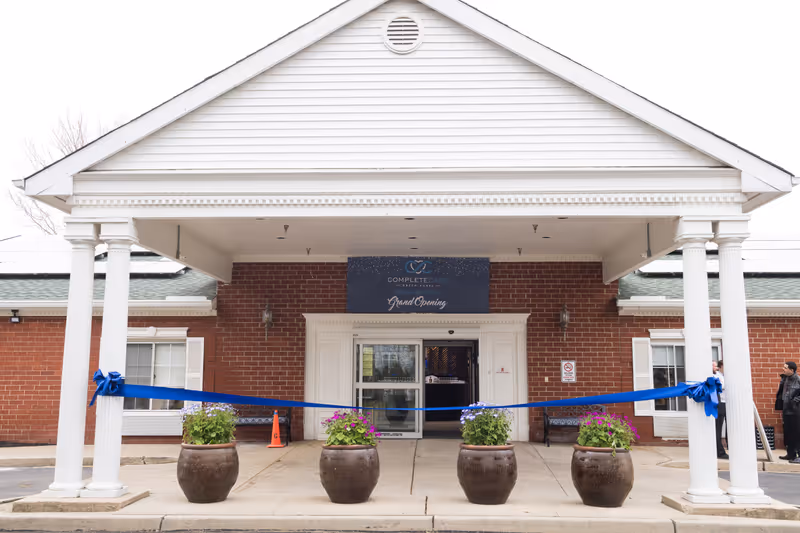 Front entrance of Complete Care at Green Acres facility with a covered porch supported by white columns. A blue ribbon is stretched across the entrance, tied to the columns, indicating a grand opening event. Four large flower pots with blooming flowers are placed in front of the entrance. Two people are standing near the right side of the building.