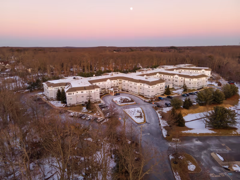Aerial view of Monarch Bella Terra facility surrounded by trees with some snow on the ground and rooftops during sunset or sunrise, with a clear sky and a visible moon.
