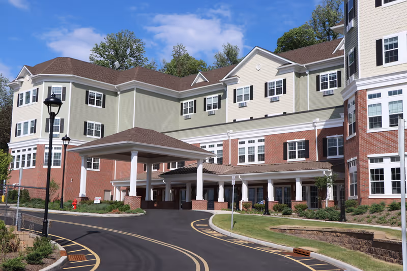 Exterior view of a multi-story assisted living facility building with a covered entrance driveway, brick and light green siding, multiple windows with black shutters, street lamps, and landscaped greenery under a partly cloudy blue sky.