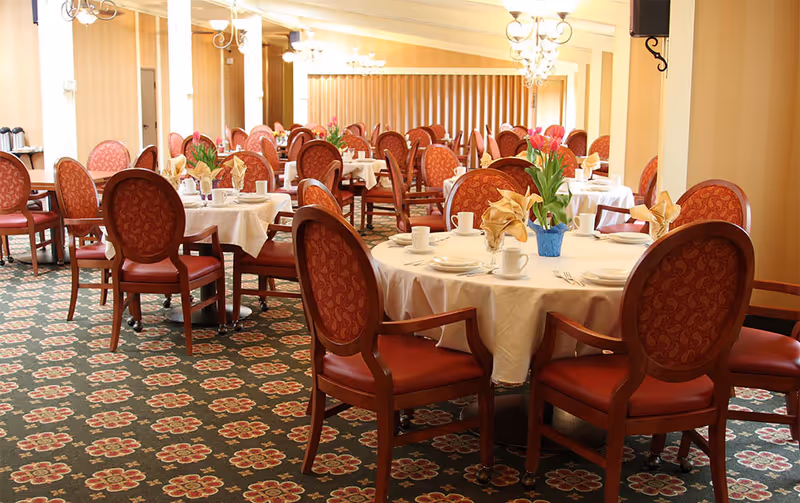 Dining room with round tables set with white tablecloths, red upholstered chairs, and floral centerpieces.