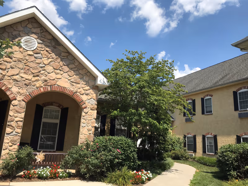 Exterior view of a senior living facility with a stone and brick facade on the left building and a beige stucco building with black shutters on the right. There is a curved concrete walkway surrounded by green bushes, flowering plants, and a tree under a blue sky with scattered clouds.