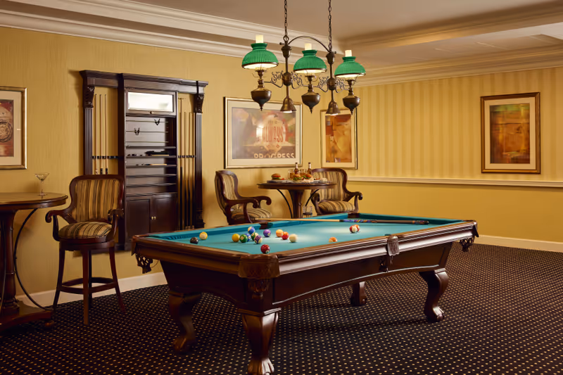 A well-appointed interior game room with a pool table, green-shaded overhead lights, and seating against yellow wallpapered walls.