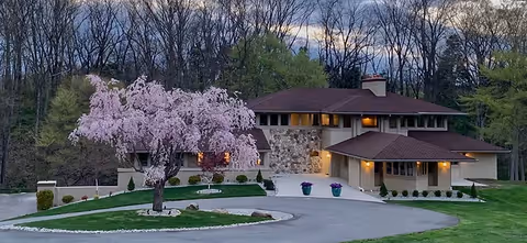 A large, modern two-story house with a stone and beige exterior surrounded by a curved driveway and well-maintained lawn. A blooming pink tree is prominently in front of the house, with a wooded area in the background under a cloudy sky at dusk.