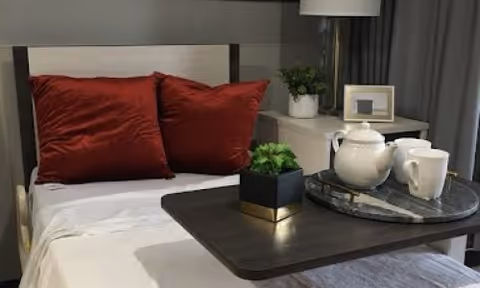 A neatly made single bed with two red pillows, an overbed table holding a teapot, mug and small plant, and a bedside table with a lamp and framed photo.