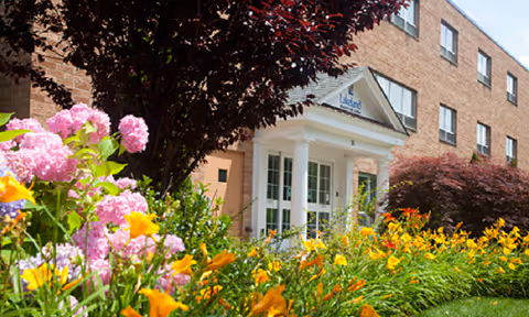 Entrance of Lakeland Health Care Center with a brick exterior, white columns, and a garden full of colorful flowers including pink, yellow, and orange blooms in the foreground.