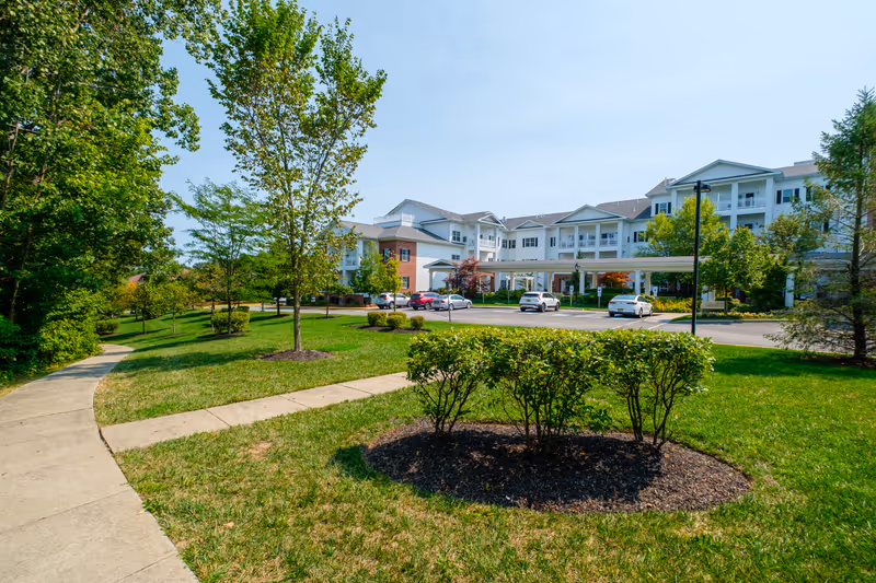 Exterior view of Brightview Randolph senior living facility showing a large white multi-story building with balconies, surrounded by green lawns, trees, and shrubs under a clear blue sky. A paved walkway curves through the landscaped grounds, and several cars are parked near the building entrance.