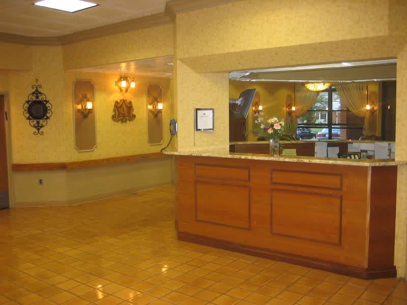 Reception area with a wooden front desk and a large mirror behind it reflecting a chandelier and windows with curtains. The walls are painted yellow with decorative sconces and wall art. The floor is covered with yellow tiles.