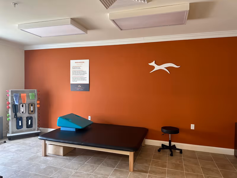 A therapy or rehabilitation room with a black padded treatment table, a teal wedge cushion on top, a black rolling stool, and a rack holding various exercise bands and weights. The room has a burnt orange accent wall with a white fox logo and a mission statement poster.