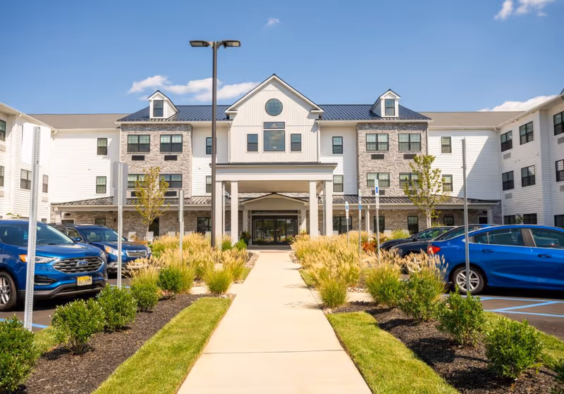 Front exterior view of a three-story senior living facility with a covered entrance, landscaped walkway, and parked cars on either side under a clear blue sky.