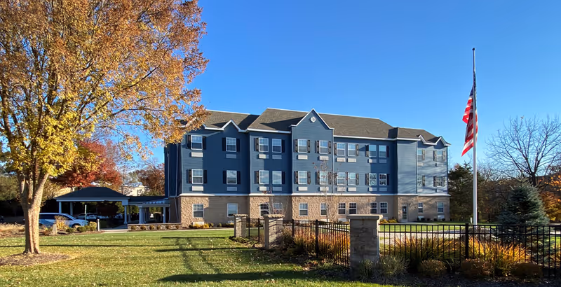 Exterior view of a three-story assisted living facility building with blue siding and beige stone accents. The building is surrounded by a well-maintained lawn, a black metal fence, and trees with autumn foliage. An American flag is flying at half-staff on a flagpole in front of the building under a clear blue sky.