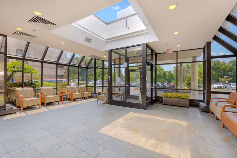 A bright and spacious lobby area with large glass windows and a glass door entrance. The room features a skylight allowing natural light to flood the space. There are several cushioned chairs with wooden armrests arranged along the walls, small tables with plants, and a clock mounted on the glass door frame. Outside, cars and greenery are visible through the windows.