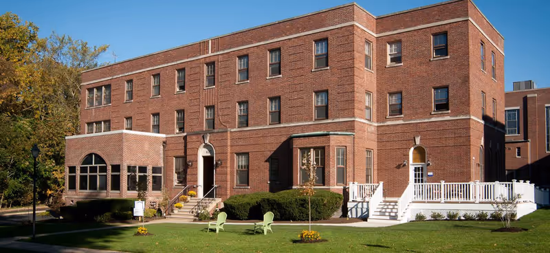 Three-story red brick senior residence building with white entryways, a green lawn and a couple of chairs in front under a clear blue sky.