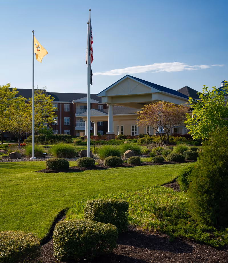 Exterior view of Applewood Village senior living facility showing a well-maintained lawn with trimmed bushes and trees, a covered entrance supported by white columns, and three flagpoles with flags flying against a clear blue sky.