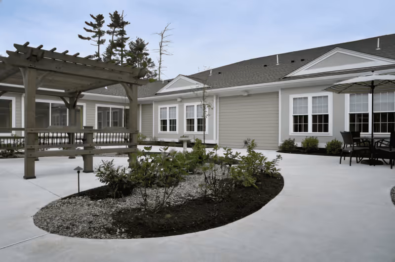 Courtyard with a wooden pergola, planted beds, outdoor seating, and the exterior of a single-story senior living building.
