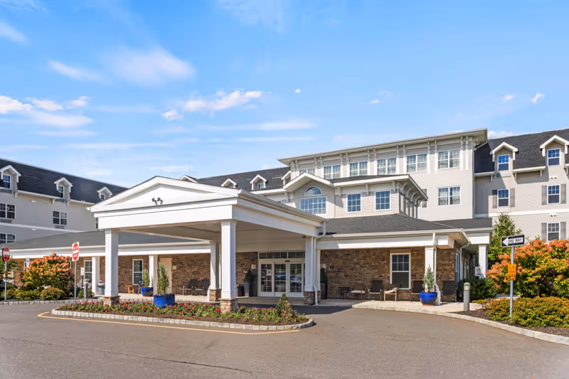 Front exterior view of a senior living facility with a covered entrance, stone and siding facade, multiple windows, and landscaped flower beds under a blue sky.