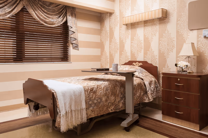 A neatly made single bed with a floral patterned bedspread and a white throw blanket at the foot. A small overbed table holds a mug and a book. Next to the bed is a wooden nightstand with three drawers, a lamp, and a small flower arrangement. The room has beige and cream striped wallpaper with a damask pattern and a window with wooden blinds and decorative curtains.