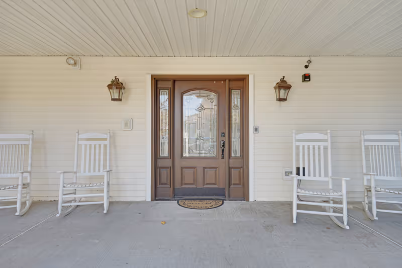 Front entrance of a building with a wooden door featuring decorative glass panels. The porch area has a white ceiling and siding, with four white rocking chairs evenly spaced on either side of the door. Two lantern-style wall lights are mounted on the wall beside the door.