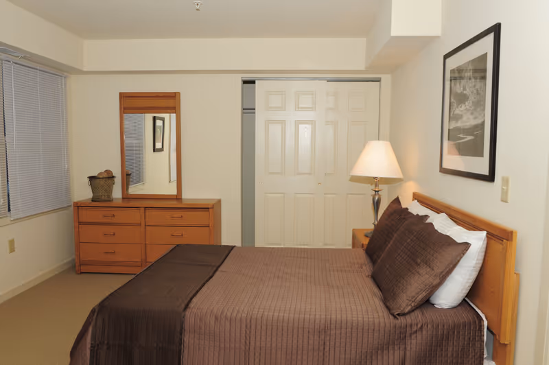 A bedroom with a neatly made bed featuring brown bedding and pillows. To the right of the bed is a wooden nightstand with a lamp. On the wall above the bed is a framed black and white picture. Across from the bed is a wooden dresser with a mirror and a decorative basket on top. There is a window with closed blinds on the left wall and a closet with sliding doors in the center background.