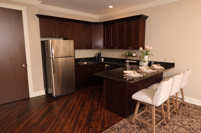 A modern kitchen with dark wood cabinets and a stainless steel refrigerator. The kitchen features a black granite countertop with a breakfast bar set with four beige upholstered chairs. The countertop is decorated with plates, napkins, glasses, and a vase with flowers. The floor is a combination of dark wood and patterned carpet.