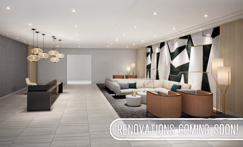 Modern and spacious lobby area with a reception desk on the left, featuring hanging pendant lights above. On the right, there is a seating area with a large white sectional sofa, two brown armchairs, round coffee tables, and floor lamps. The back wall has a geometric art installation. The floor is tiled, and the ceiling has recessed lighting.