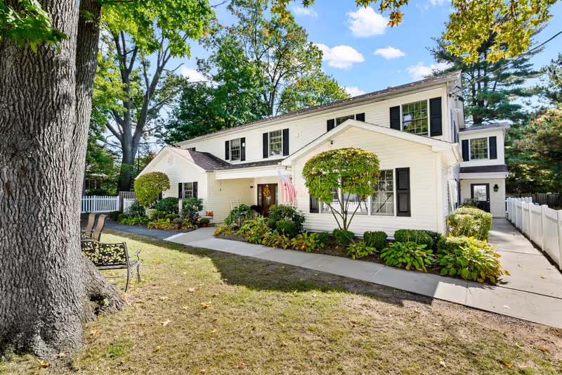 A two-story white building with black shutters surrounded by green trees and shrubs under a blue sky with scattered clouds. There is a concrete walkway leading to the entrance, which has an American flag displayed. A large tree and a bench are visible in the foreground on a grassy area.