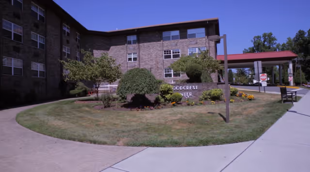 Exterior view of a multi-story brick building with several windows, surrounded by a landscaped garden with trimmed bushes and flowers. A curved sidewalk leads to the building entrance, which has a covered drop-off area with a red roof. There are signs near the entrance and a bench along the sidewalk.