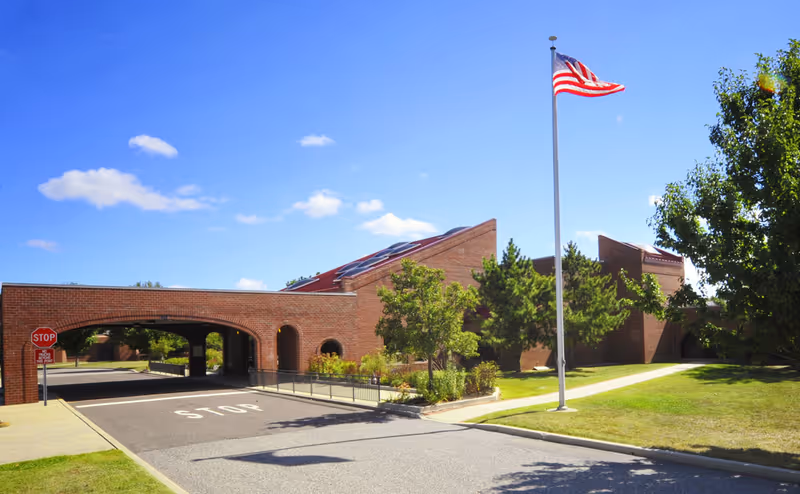 Exterior view of a brick building with an arched driveway, a stop sign, a flagpole with an American flag, and surrounding greenery under a clear blue sky.