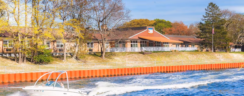Exterior view of a single-story rehabilitation and healthcare center building with a brown roof and beige walls, situated behind a grassy embankment and a red retaining wall along a body of water. A small white motorboat is seen moving on the water in the foreground, and trees with autumn foliage surround the building under a clear blue sky.