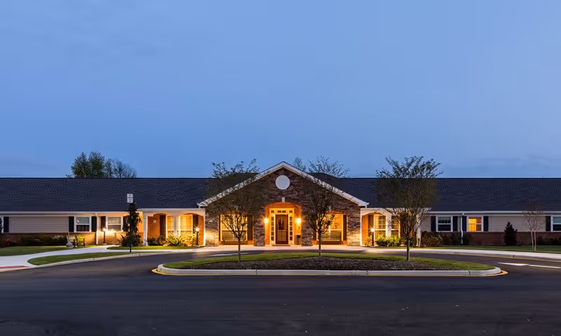 Front entrance of a single-story senior living facility at dusk with a lit entryway, circular driveway, and landscaping.