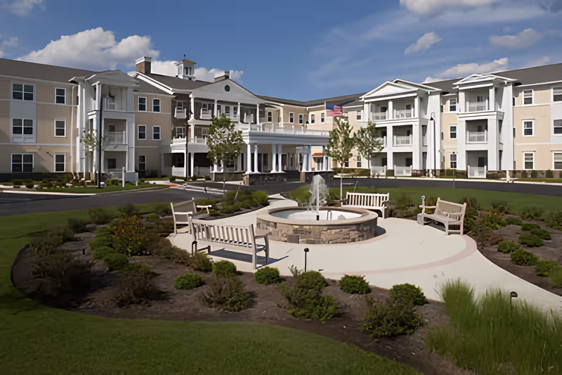 Exterior view of a senior living facility with a circular driveway, landscaped garden, benches, and a central water fountain under a partly cloudy sky.