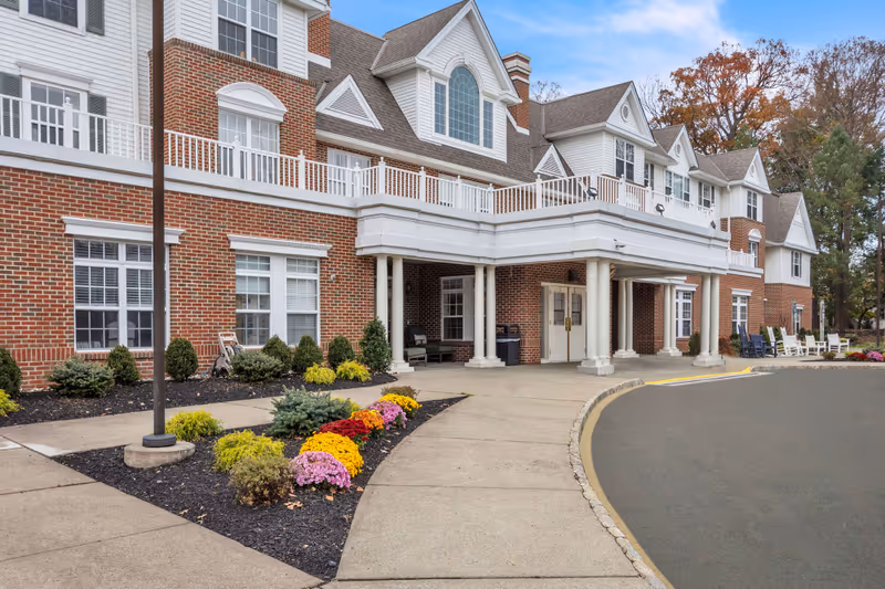 Main entrance of a red-brick senior living building with white trim, a covered drop-off portico, landscaped flower beds, and a curved driveway.