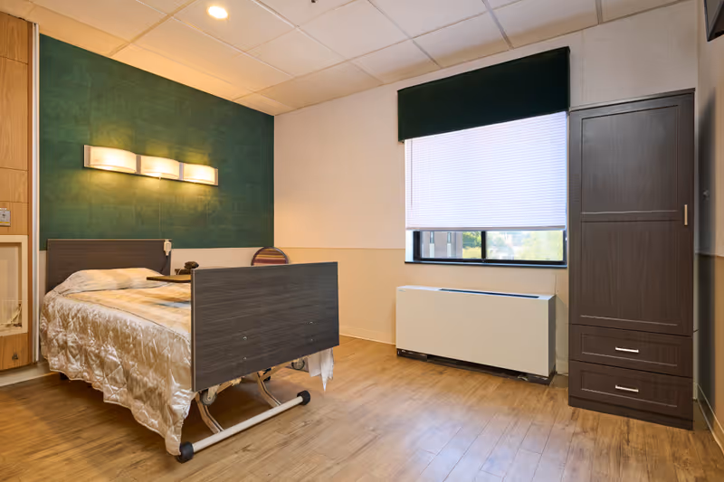 A simple, clean patient bedroom in a rehabilitation facility featuring a hospital bed with a beige quilted blanket, a wooden cabinet, a chair, a window with a white blind and dark green valance, and a green accent wall with a modern wall light fixture above the bed.