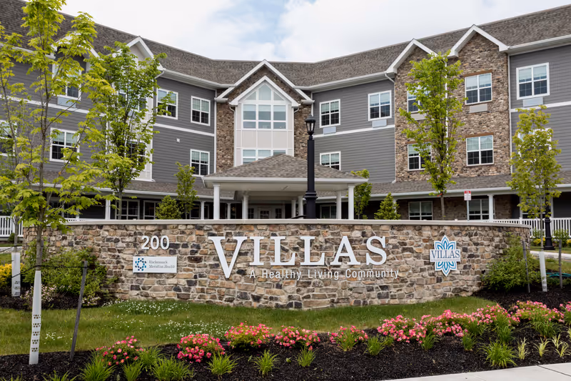 Stone sign reading 'VILLAS' in front of a three-story senior living building with landscaping.