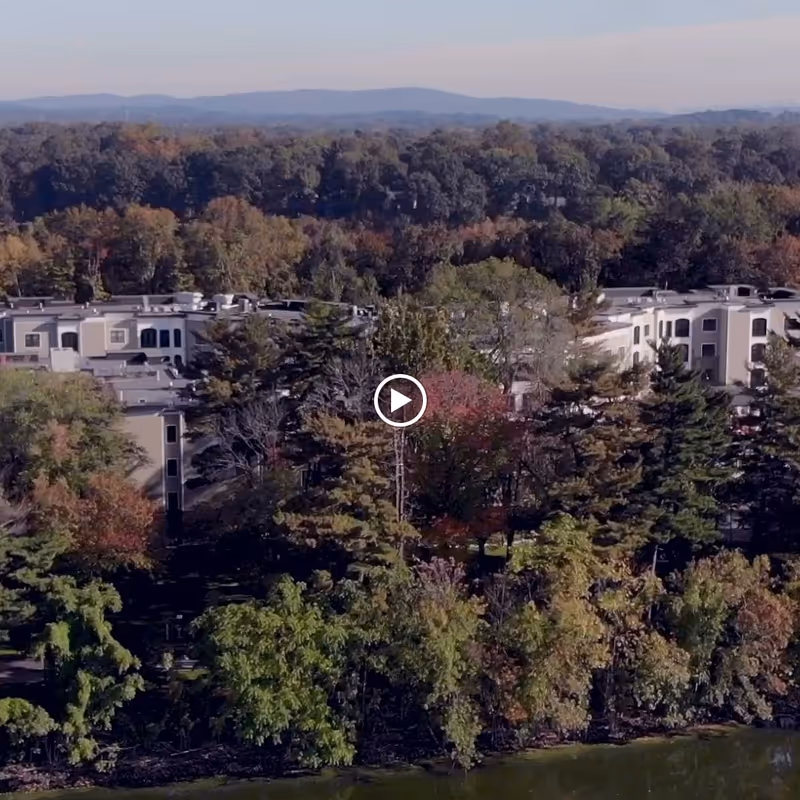 Aerial view of Allegro Senior Living facility in Harrington Park, NJ, surrounded by dense trees with autumn foliage and a body of water in the foreground, with hills visible in the distance under a clear sky.
