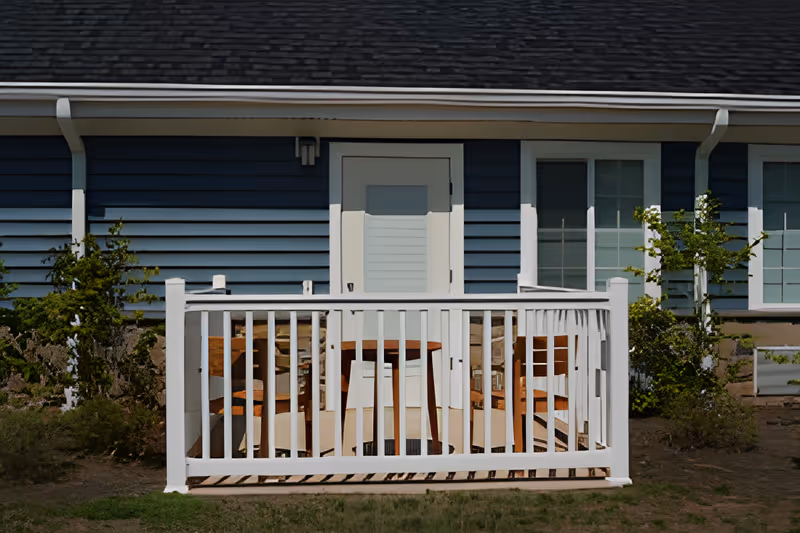 Small enclosed porch with a white railing, wooden chairs and table in front of a blue-sided building with a door and windows.