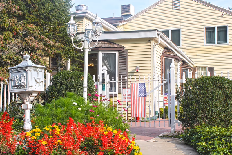 Exterior view of a yellow senior living facility building with white trim, surrounded by a white picket fence and colorful flower beds with red, yellow, and pink flowers. There is a decorative white mailbox and a vintage-style street lamp near the entrance. An American flag is displayed on the fence.