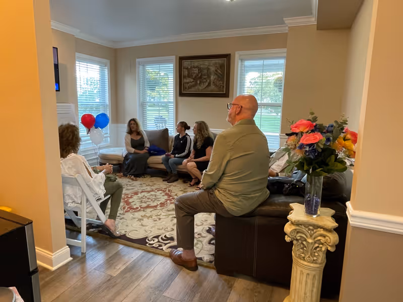 A group of people seated in a bright living room with balloons, a patterned rug, and a vase of flowers.