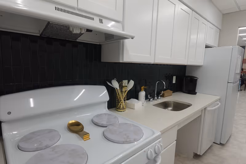 A clean and modern kitchen area with white cabinets, a white stove with marble-patterned burner covers, a small sink with a faucet, a soap dispenser, a utensil holder with white utensils, a coffee maker, and a white refrigerator. The backsplash is made of dark tiles, and the floor is light-colored.