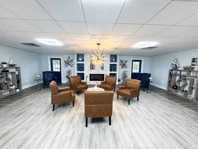 A spacious and modern living room with light wood flooring and a white ceiling with recessed lighting. The room features a central seating area with five brown leather armchairs arranged around a small round table with a vase of pink flowers. On either side of the room, there are blue armchairs near dark blue doors with windows. The back wall has a decorative fireplace surrounded by white and blue shelving units displaying various decorative items and artwork. Two wooden shelving units with nautical-themed decor are positioned against the side walls.