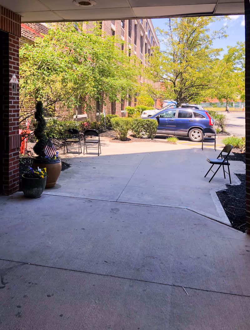 Covered entrance and driveway of a brick residential care building with potted plants, folding chairs, landscaping, and a parked blue car.