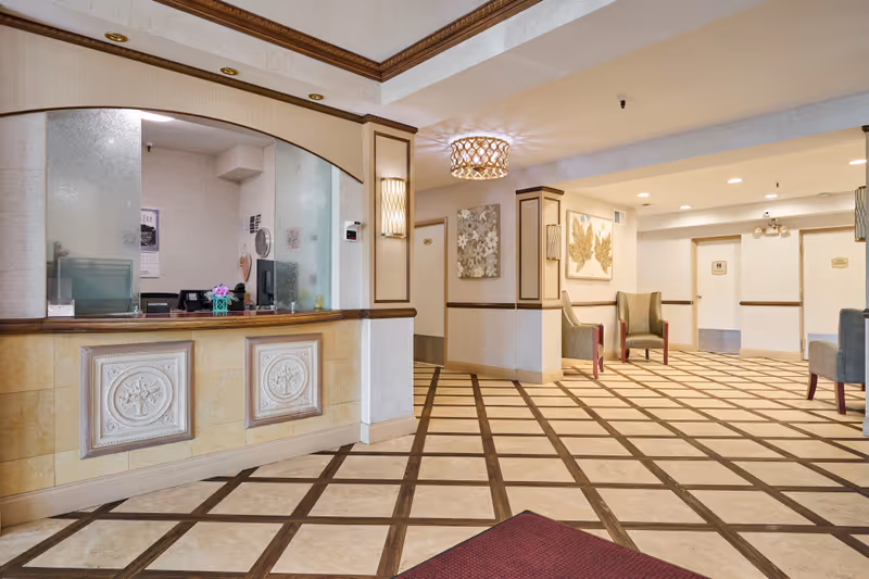 Interior view of a senior living facility reception area with a front desk on the left, decorative wall panels, patterned tile floor, and several chairs along the walls. The area is well-lit with ceiling lights and wall sconces, and there are framed artworks on the walls.