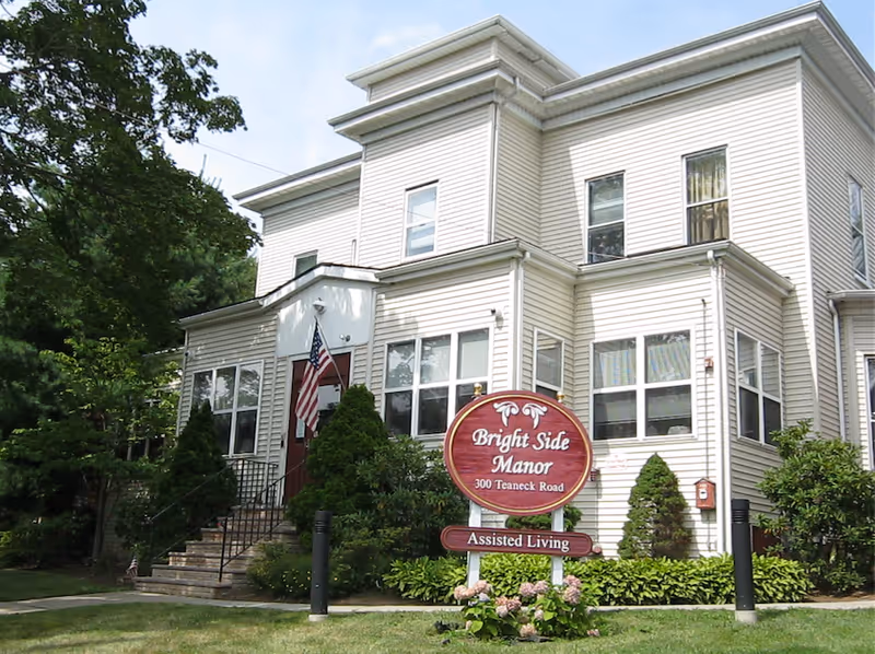 Front exterior of a beige two-story assisted living building with an American flag and a 'Bright Side Manor' sign on the lawn.