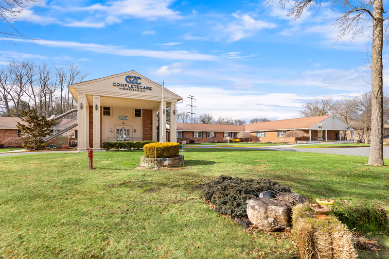 Front exterior of CompleteCare at Mercerville showing the building entrance with columns, a lawn, and adjoining single-story wings under a blue sky.