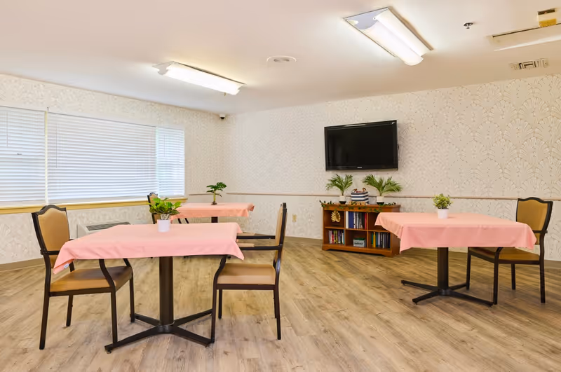 A small dining area with three tables covered in pink tablecloths, each with a small potted plant on top. There are chairs around the tables, a wooden bookshelf with books and plants, and a flat-screen TV mounted on a wallpapered wall. The room has large windows with blinds and wood-look flooring.