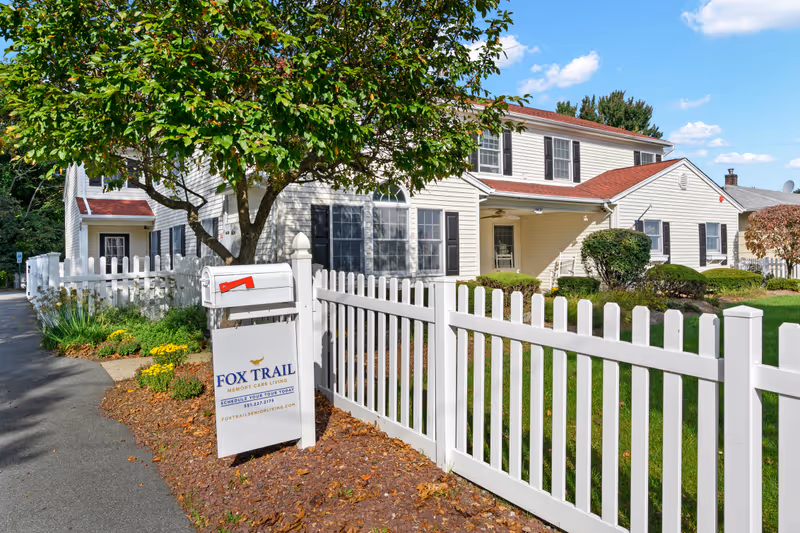 Exterior view of Fox Trail Memory Care at Paramus, showing a white two-story building with black shutters and a red roof. A white picket fence runs along the front yard with green grass and bushes. A white mailbox and a sign with the facility's name and contact information are visible near the sidewalk. A tree with green leaves partially shades the area.