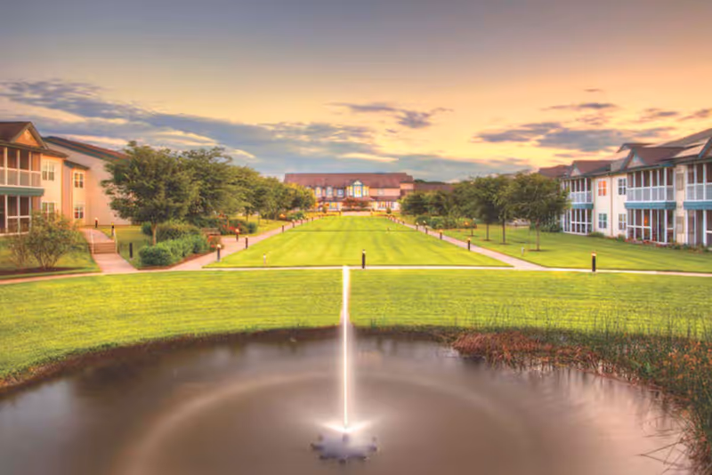 A landscaped outdoor area at Stonebridge at Montgomery Healthcare Center featuring a central water fountain in a pond, flanked by green lawns and trees, with two residential buildings on either side and a larger building in the background under a colorful sunset sky.